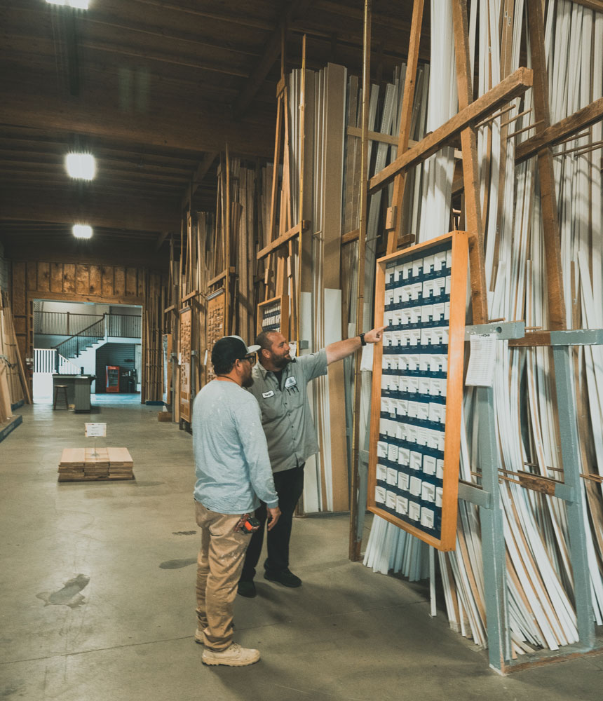 Two men in a warehouse, with one pointing at a large board filled with small labeled boxes of mdf molding while discussing.